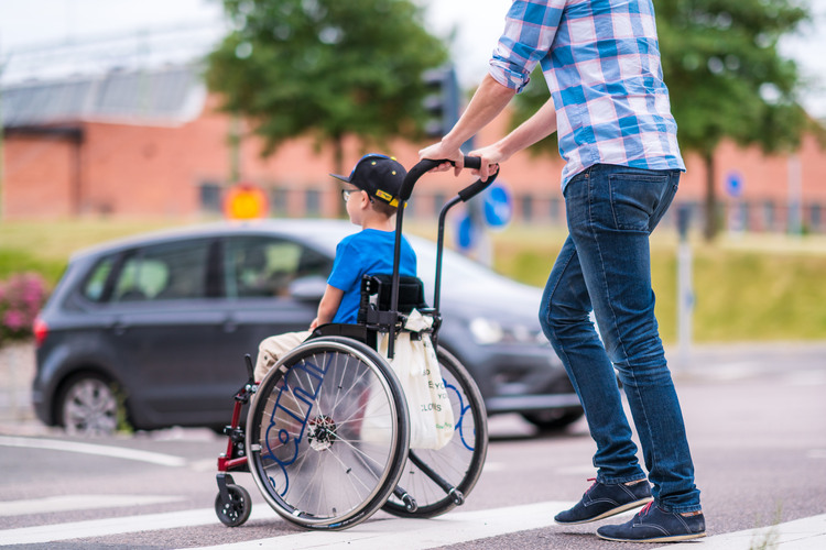 Ett barn som sitter på en rullstol och en vuxen som hjälper med att köra den 