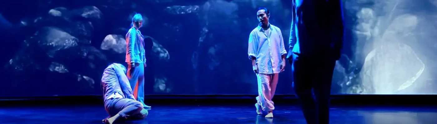 Skådespelare på en scen som badar i blått ljus mot en bakgrund av vatten